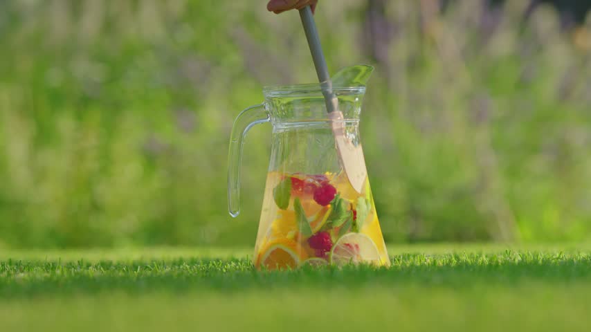 Hand mixes cold citrus fruit drink with raspberries in transparent jug sitting outdoors on lush grass. Summer picnic beverage preparation captured in bright cinematic footage.