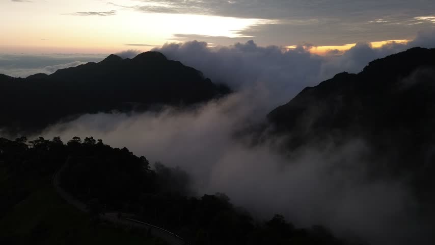 A misty mountain ridge at dusk with rolling cloud, forested valley, layered peak landscape, glowing sunset horizon, dramatic highland scenery, tropical slopes and atmospheric alpine view