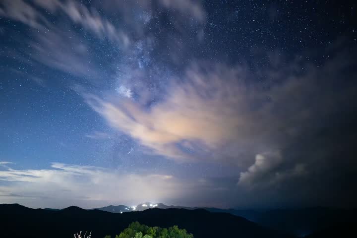 A night mountain landscape under glowing cloud and starry sky, milky way above rugged ridge, distant light across peaceful wilderness, tranquil starlight scene
