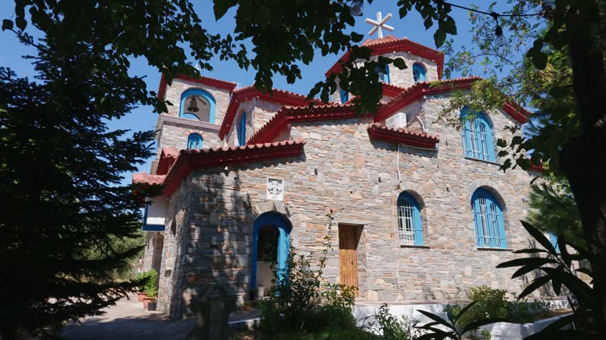 Traditional Greek Orthodox church with white stone walls and red tiled roofs surrounded by mature trees on the island of Thassos, Greece. Static travel footage showing Mediterranean religious architec