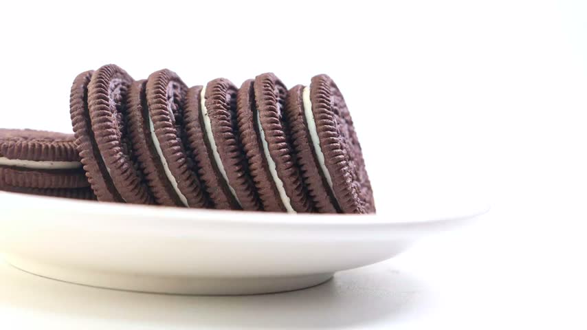 Stacked chocolate sandwich cookies with white cream filling resting on white plate against clean white background. High quality close up footage perfect for food content creation.