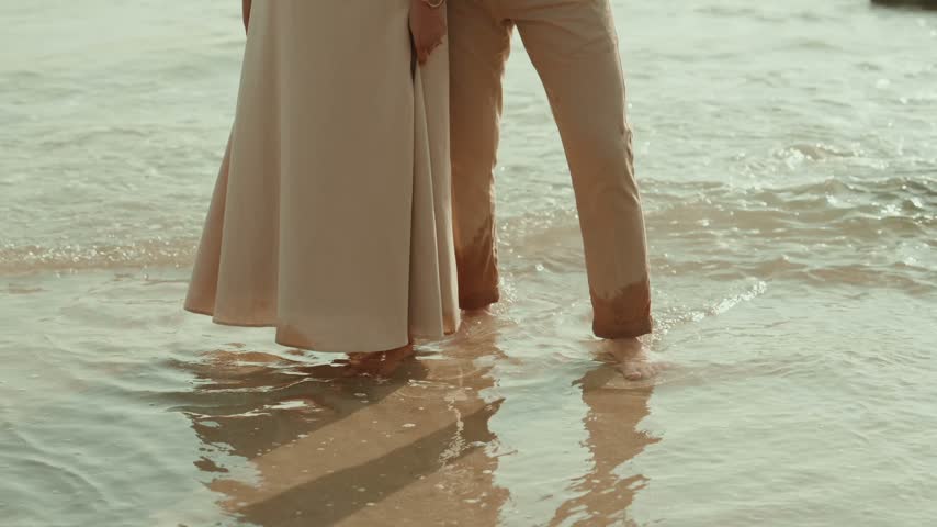 Couple stands barefoot in shallow water at the shoreline, his hand resting on her waist, both wearing beige.