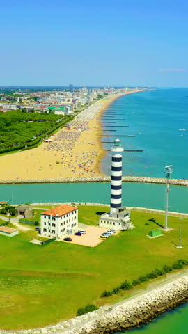 Vertical video. Italy, Jesolo. The Piave Vecchia lighthouse, a 48-metre cylindrical tower painted with bold black and white horizontal bands, stands at the mouth of the Sile River where it meets the Adriatic Sea. Originally built in 1846 under Austrian rule and rebuilt between 1949 and 1951 after its destruction during the Second World War, the lighthouse features a spiral staircase of 243 Istrian stone steps and a light visible up to 18 nautical miles away. The aerial drone footage captures the tower from an elevated perspective, sweeping forward over the river channel to reveal the expansive sandy beach stretching along the coastline. Dense green pine forests line the inland edge of the shore, while rows of beach umbrellas and sunbathers dot the golden sand below. The turquoise Adriatic water transitions from shallow green near the shore to deep blue further out, with small boats scattered across the calm surface. A long stone breakwater extends into the sea, paralleled by wooden groynes that segment the beach at regular intervals. The city of Jesolo spreads along the horizon under a clear, cloudless summer sky, with hotels and residential buildings visible in the distance. The scene conveys a vibrant, sunlit atmosphere of coastal leisure and maritime heritage. Aerial View. Rich colors