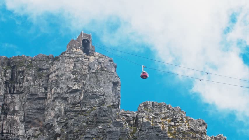 Tracking shot of a Table Mountain cable car ascending a rugged rock face against a clear blue sky, Cape Town, South Africa