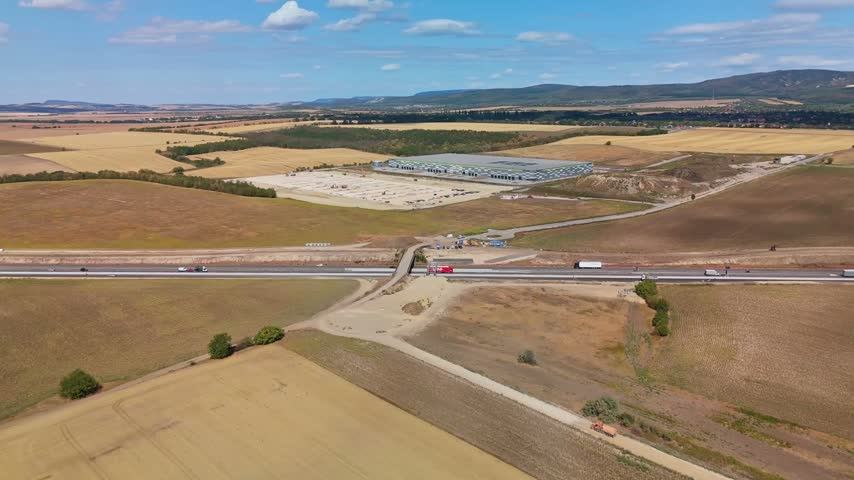 Drone view of M1 highway construction beside a new logistics warehouse and agricultural fields near Páty, Hungary, with freight traffic moving through the rural landscape.