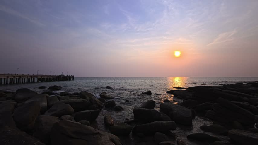 Tropical island sunset over the sea on Koh Kood, Thailand, with sun reflections on ocean water. This paradise island in Southeast Asia has an orange sunset sky above a rocky beach and tropical coast. The sun is visible beneath clouds, with calm sea along the island shore. A concrete pier extends from a rocky coastline into the water. This tropical beach has sea waves and coastal rocks beneath a cloudy sunset sky.