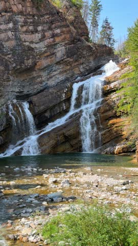 Cameron Falls cascading over layered rock cliffs into a green pool at Waterton Lakes National Park in Alberta, a scenic waterfall surrounded by trees in summer.