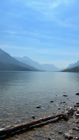 Calm clear lake with mountains and misty blue sky at Waterton Lakes National Park in Alberta, with a driftwood log and smooth stones along the pebbly shoreline.