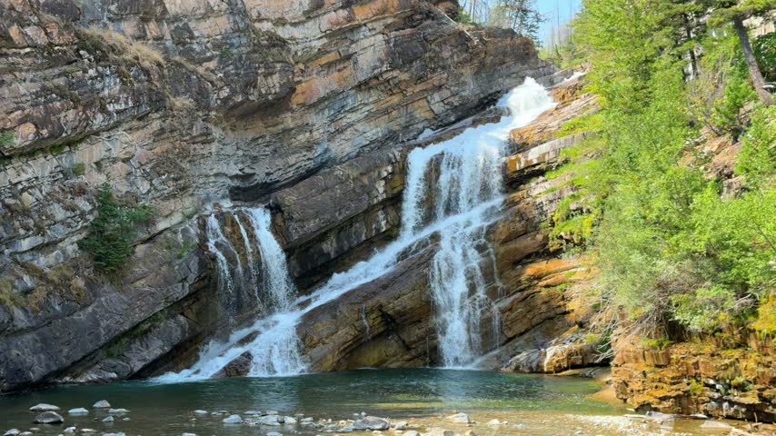 Cameron Falls cascading over layered rock cliffs into a green pool at Waterton Lakes National Park in Alberta, a scenic waterfall surrounded by trees in summer.