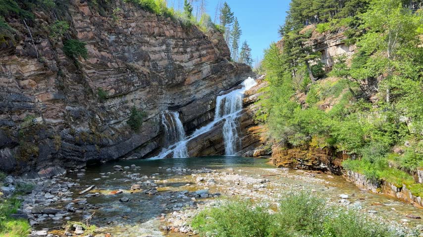 Cameron Falls cascading over layered rock cliffs into a green pool at Waterton Lakes National Park in Alberta, a scenic waterfall surrounded by trees in summer.