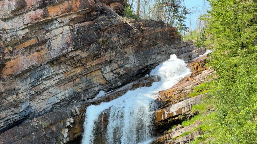 Cameron Falls cascading over layered rock cliffs into a green pool at Waterton Lakes National Park in Alberta, a scenic waterfall surrounded by trees in summer.