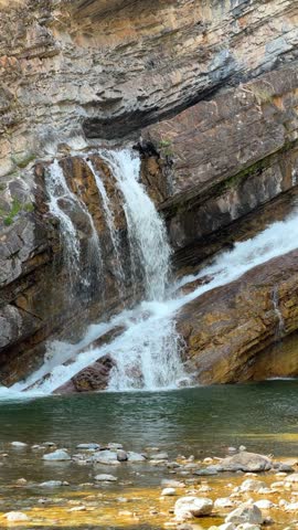 Cameron Falls cascading over layered rock cliffs into a green pool at Waterton Lakes National Park in Alberta, a scenic waterfall surrounded by trees in summer.
