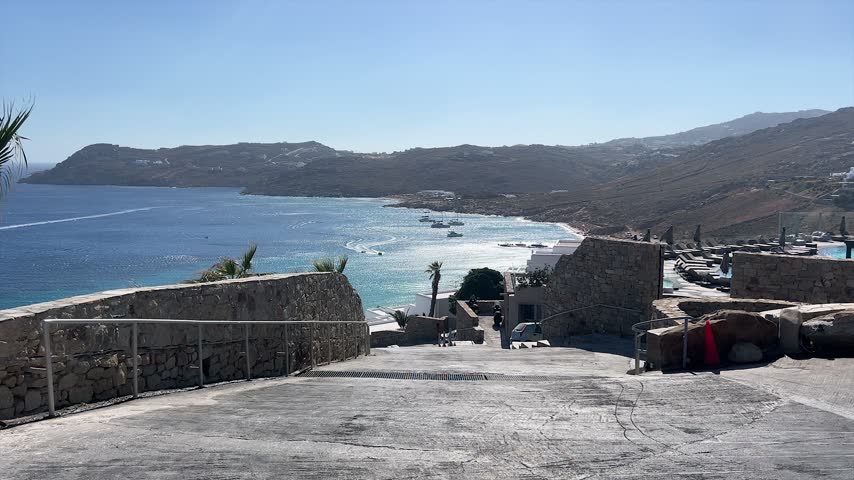 Stone walkway overlooking a bright blue bay, rocky hills, boats and seaside