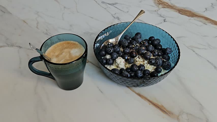 Blueberries sit in a blue bowl beside a coffee cup. Cereals and berries compose a healthy breakfast. Marble surface holds the meal setup. Spoon rests inside the bowl. The coffee mug has a spoon inside