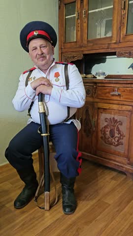 Cossack Man in historical military uniform sits indoors holding rifle. He wears cap, vest, and dark trousers with red accents. Wooden cabinet stands behind him. He rests hand on weapon stock