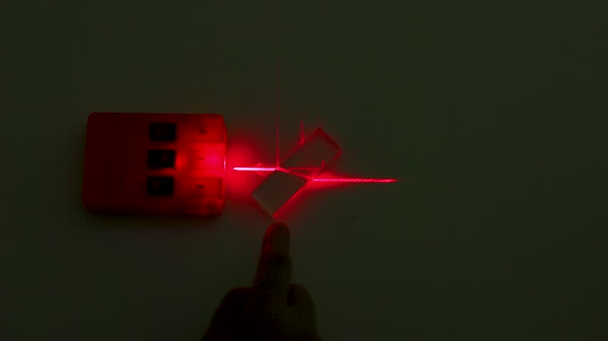 A hand adjusts an acrylic optical lens demonstrating laser beam refraction in a dark laboratory.