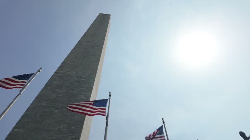 Low-angle view of the washington monument towering against a clear blue sky with the sun shining brightly, while american flags on flagpoles wave proudly in the breeze around its base