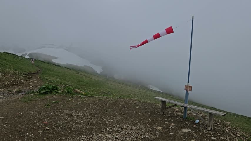 Fog blankets mountain peak with wind vane and bench. Red and white striped wind vane flaps in misty breeze. Green grass and gravel slope beneath foggy veil