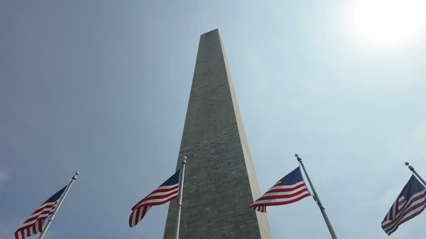 Many united states flags flying proudly in the wind at the base of the washington monument against a clear blue sky, a powerful symbol of american heritage, freedom, and national pride