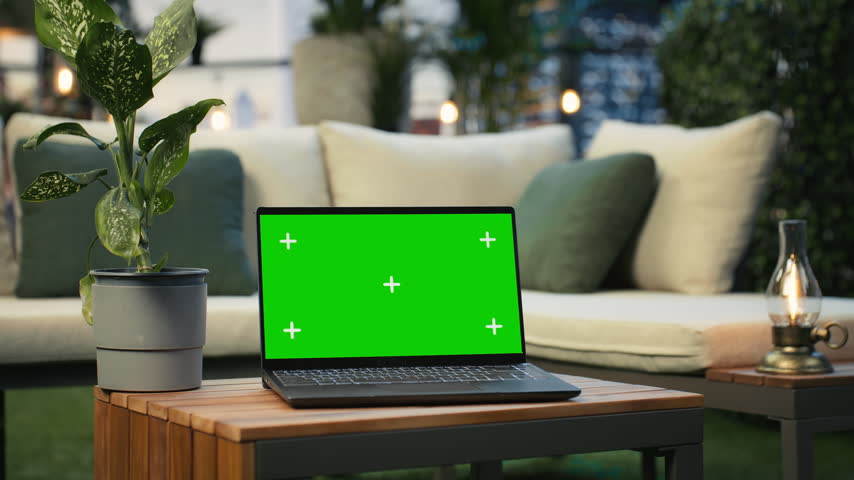 Green-screen laptop sits on a wooden table in a rooftop workspace, with leafy plants and lounge cushions behind the display