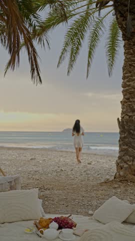 A woman walks along the beach during sunset. The sky has soft colors. There is a cozy setup on the sand with food and drinks. The sea is calm and the palm trees sway gently.