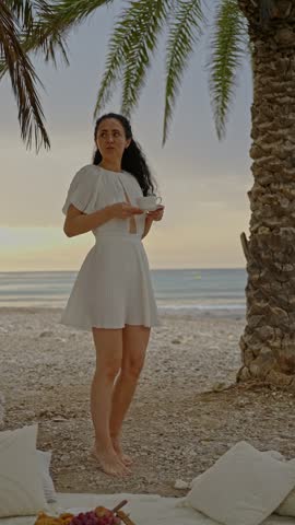 A woman is on the beach near a palm tree. She wears a white dress and holds a drink while looking towards the ocean at sunset. The scene has a relaxed atmosphere and warm colors.