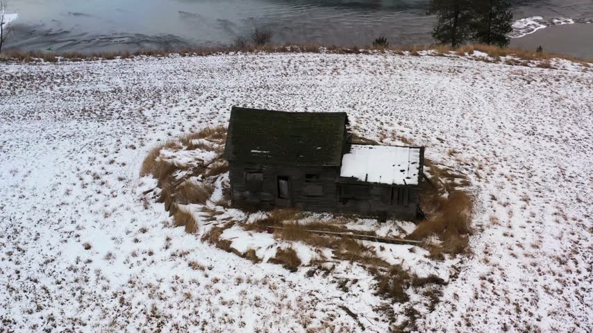 Old, weathered house rests in a snowy landscape with grass patches. Surrounding area displays patches of snow and ice near a calm river.