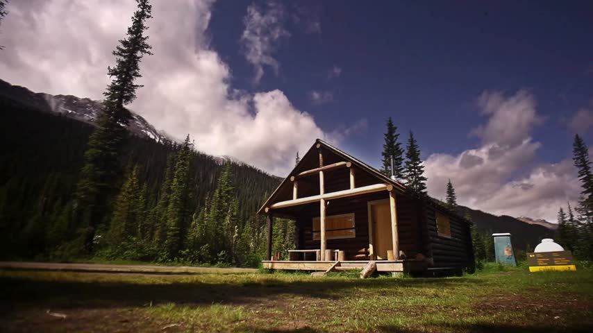 Log cabin stands in a lush green forest with mountains in the background. Pine trees surround the cabin, creating a natural and rustic environment.