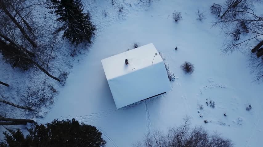 Cabin nestled in thick snow with trees surrounding it. Winter scene captures quietness and beauty of nature. Footprints lead through the snow, showcasing activity in the area.
