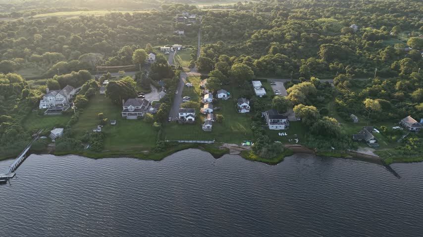 Sunlit waterfront homes and long wooden pier stretch along calm bay shoreline in Montauk, Long Island, New York, surrounded by dense green trees under golden sunrise. Aerial drone wide shot
