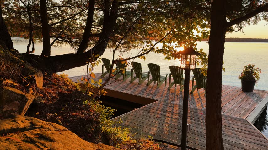 Sunset light filters through trees onto a lakeside dock with empty chairs in Muskoka
