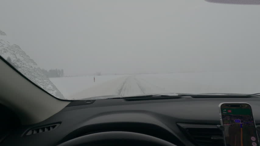 View across a car dashboard and navigation screen shows a snow-covered highway in foggy winter conditions.