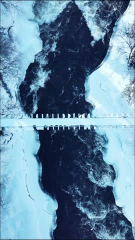 Frozen riverbanks with rushing dark water beneath a snowy landscape and wooden bridge