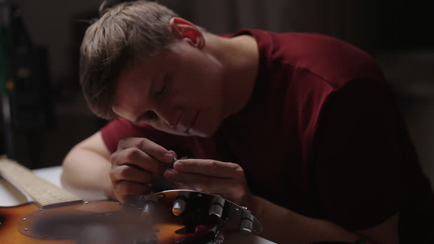 Concentrated man in red shirt leans over table, adjusting small part of electric guitar. Close-up shot showing craftsmanship and detailed musical instrument repair, setup. Shooting in slow motion.