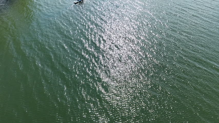 Top down drone view of a small boat moving across green lake water.