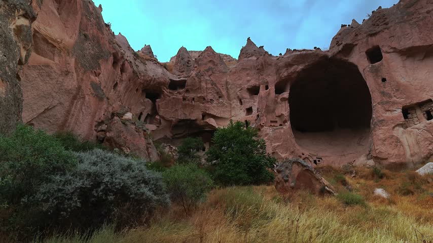Ancient cave dwellings carved into rocky cliffs with dry grass and shrubs below