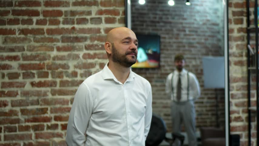 Bald man in white dress shirt speaking and reacting in loft office with exposed brick and blurred colleague