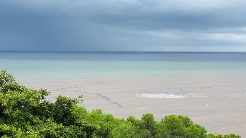 Muddy brown sediment water from heavy rain runoff mixing with turquoise Mediterranean sea under dark storm clouds, waves breaking on the shore, aerial view from hill with pine trees