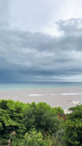 Muddy brown sediment water from heavy rain runoff mixing with turquoise Mediterranean sea under dark storm clouds, waves breaking on the shore, aerial view from hill with pine trees