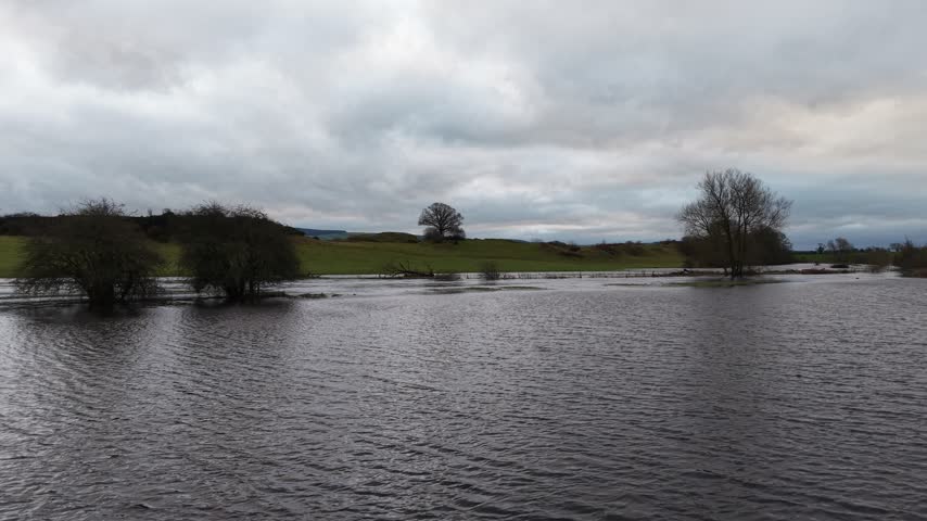 Wide view of a flooded rural meadow with leafless trees, calm water surface, and heavy overcast clouds above green countryside. Natural landscape scene showing seasonal flooding after rain.