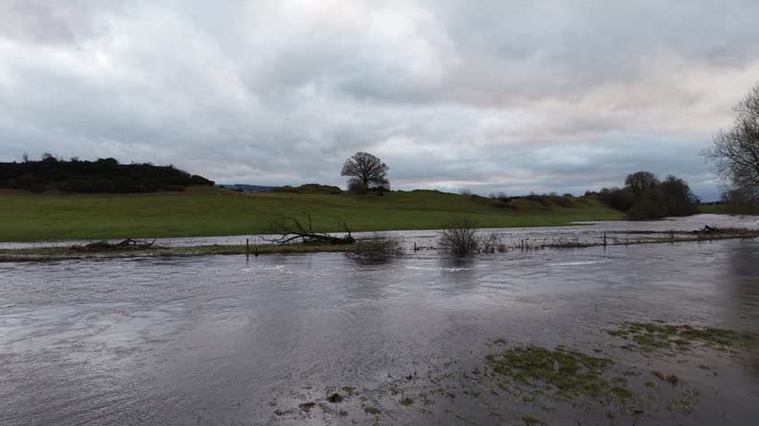 Wide view of a flooded rural meadow with leafless trees, calm water surface, and heavy overcast clouds above green countryside. Natural landscape scene showing seasonal flooding after rain.