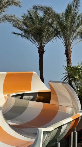 A bright orange and white water slide stands at a water park surrounded by palm trees. The sun shines down as guests enjoy the attractions in the afternoon.
