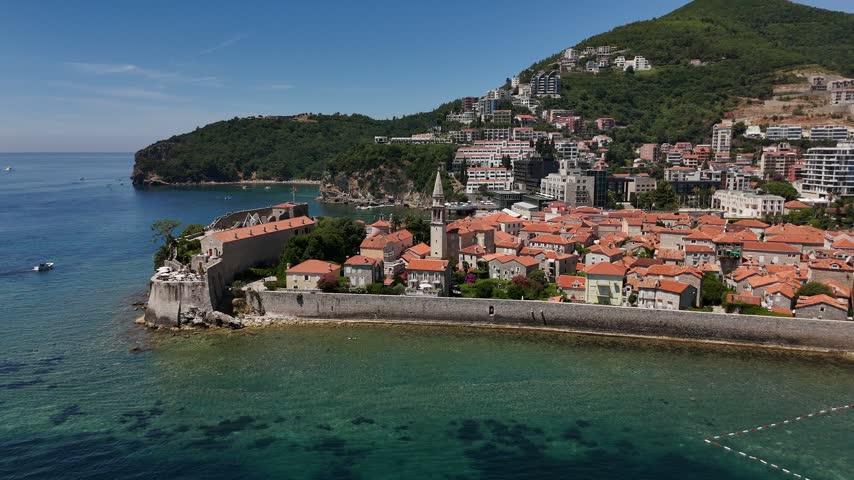 High-angle aerial panorama of Budva Old Town in Montenegro with ancient stone walls, red-roofed houses, coastal hotels, green hills and turquoise Adriatic water.
