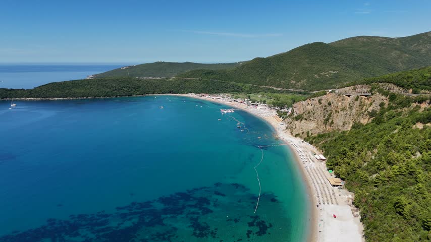 Aerial panoramic view of Jaz Beach in Montenegro, showing a long sandy coastline, umbrellas, clear turquoise Adriatic water and green hills on a sunny day.