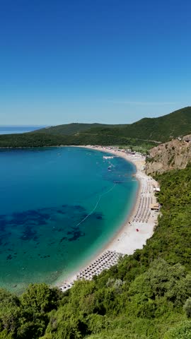 Vertical aerial view of Jaz Beach in Montenegro with a long sandy shoreline, colorful umbrellas, crystal-clear turquoise water and lush green hills under blue skies.