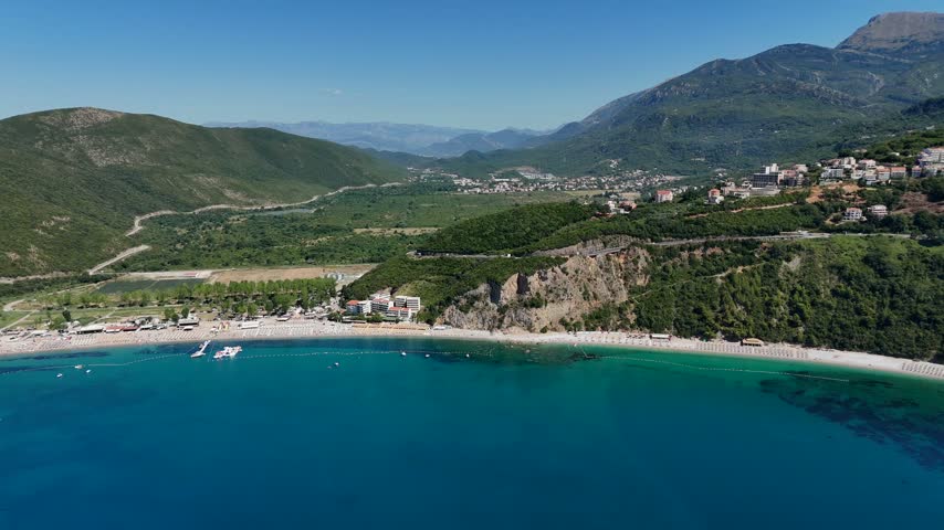 High-angle aerial view of Jaz Beach in Montenegro with golden sand, sunbeds, clear Adriatic water, coastal facilities and a massive green mountain backdrop.
