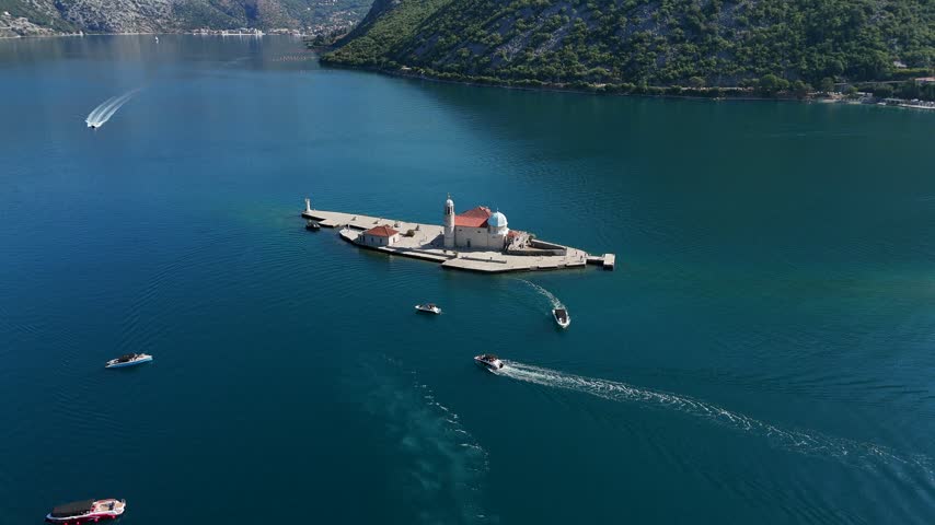 Top-down drone view of Our Lady of the Rocks artificial island and historic church in Montenegro, surrounded by deep blue water with small boats nearby.