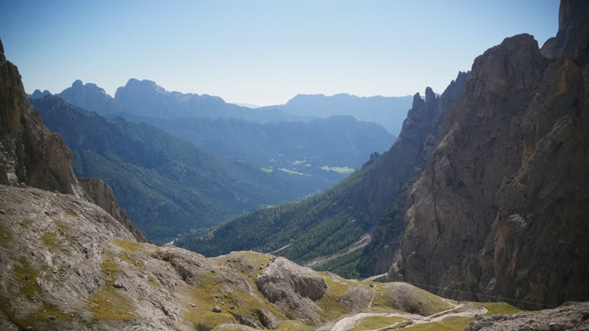 Rugged high pass trail winds between steep rock walls toward layered forest hills and distant ridges in late summer daylight.