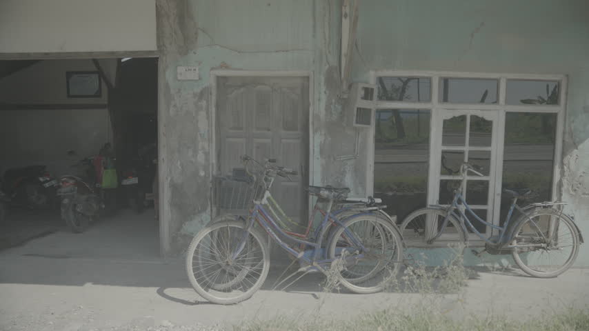 Ungraded log footage of vintage bicycles leaning against an old weathered building with peeling turquoise paint, a wooden door and white framed windows, with motorbikes parked in a doorway, flat color profile for color grading