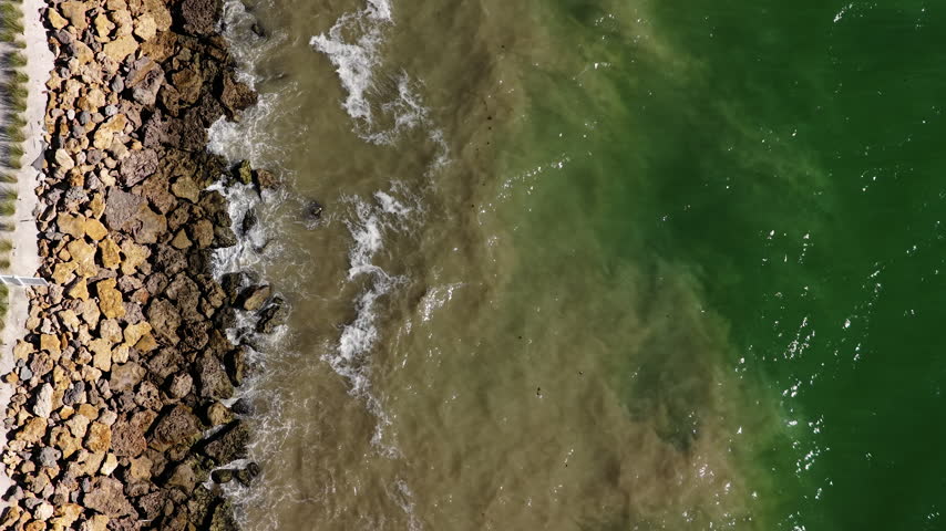 Waves wash over rocks along a coastal breakwater, where green seawater meets a band of murky water beside a narrow path.